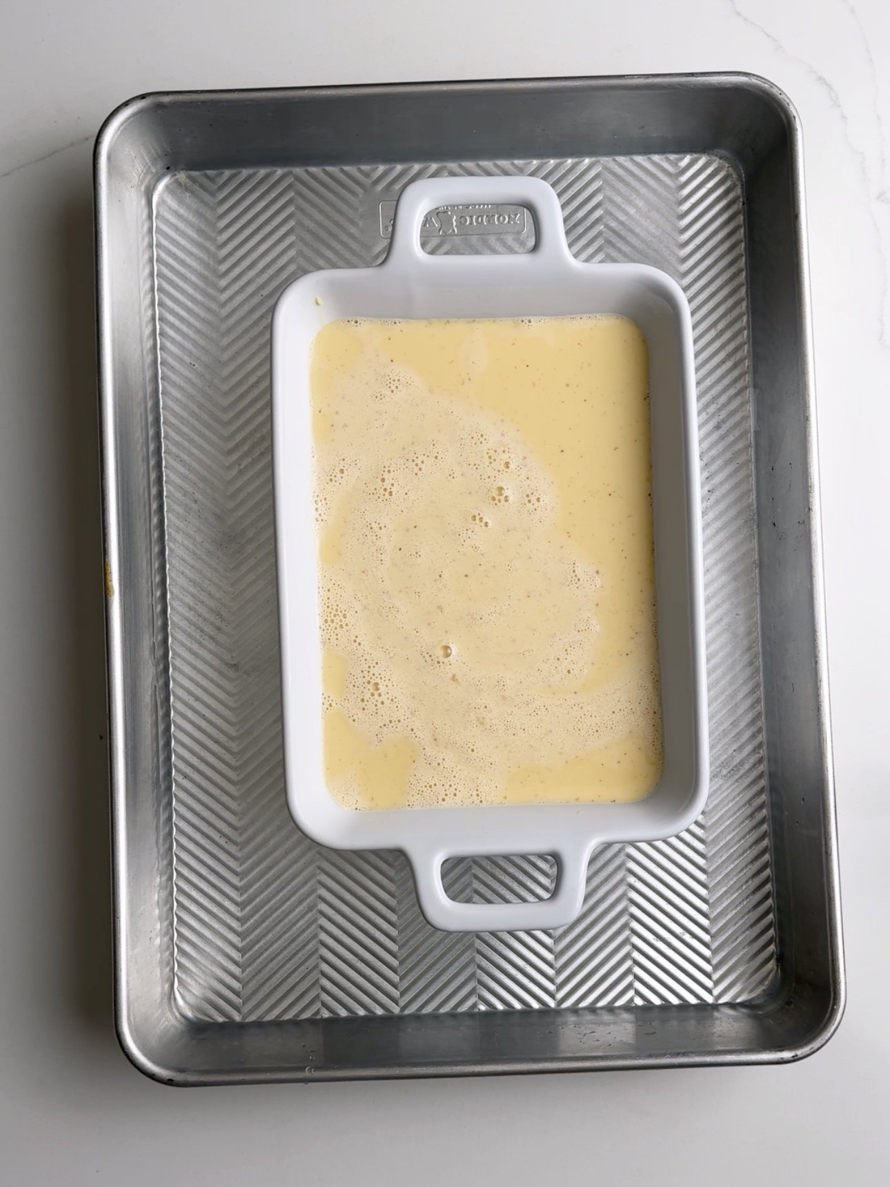 Custard poured into a white rectangular baking dish. The baking dish is in a rimmed sheet pan.