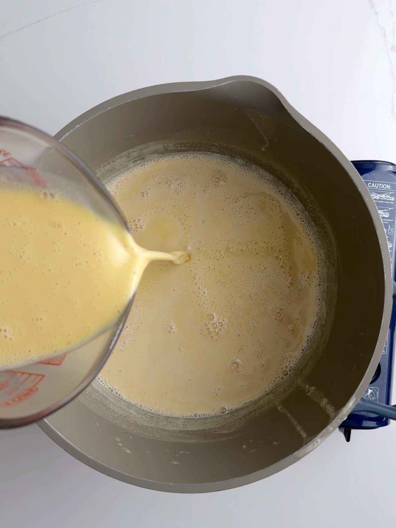 Pouring tempered eggs into a pot of warm milk to make baked custard