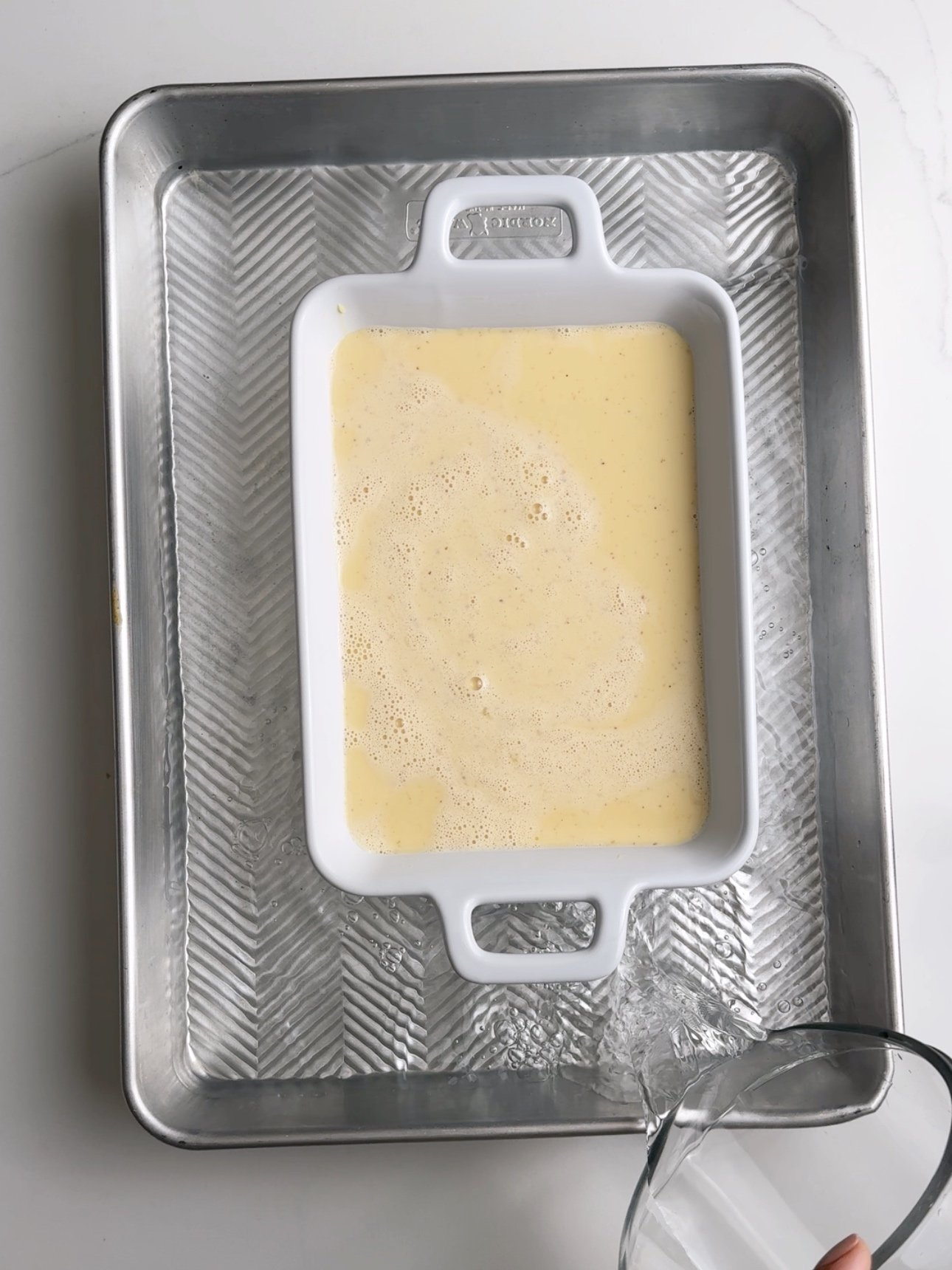 Custard poured into a white rectangular baking dish. The baking dish is a a rimmed sheet pan filled with water to create a bain-marie.