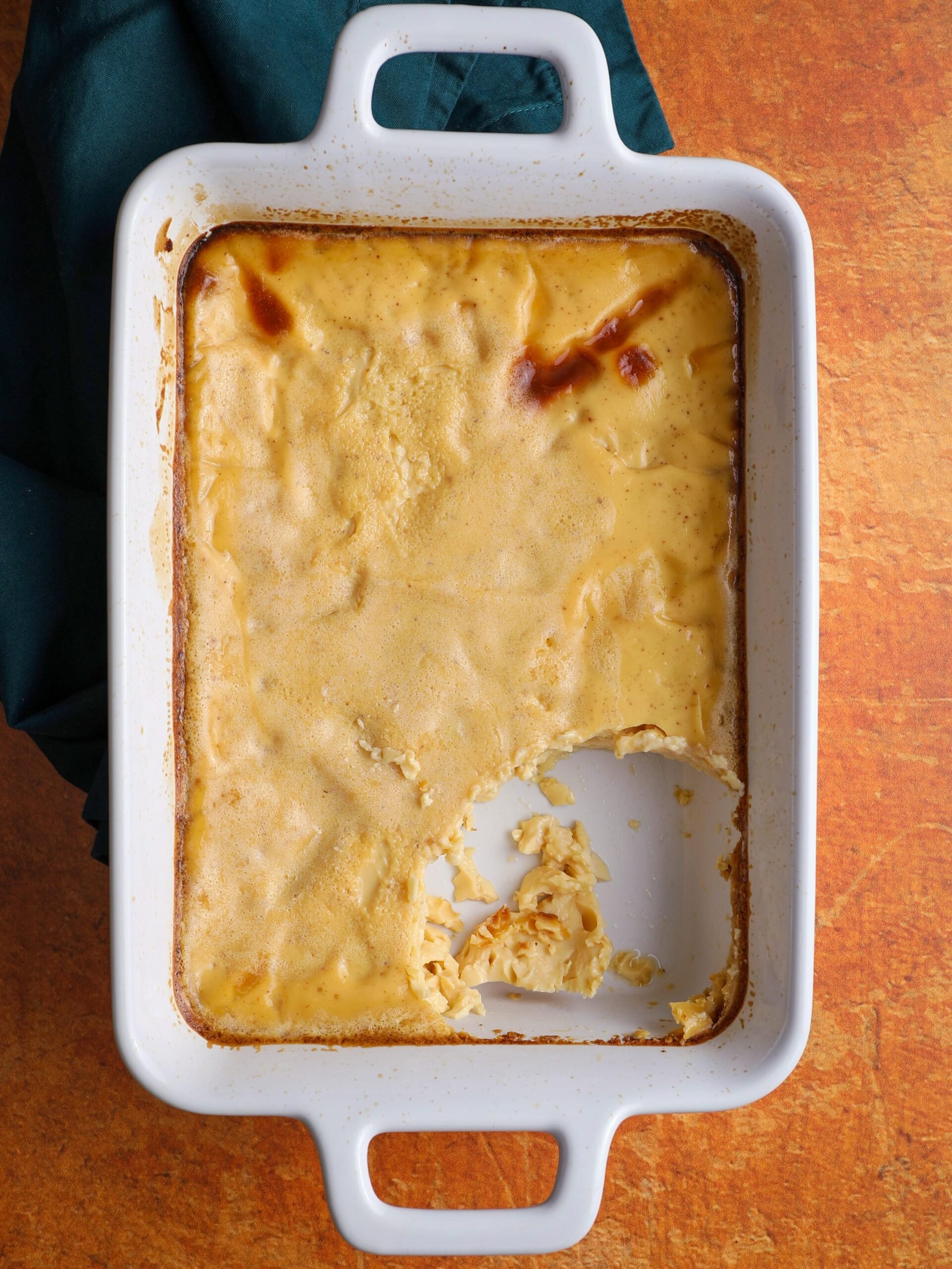 Baked custard in a white rectangular dish with handles on both ends on an orange colored background and a green napkin on the left. A large scoop of baked custard is missing from the dish.