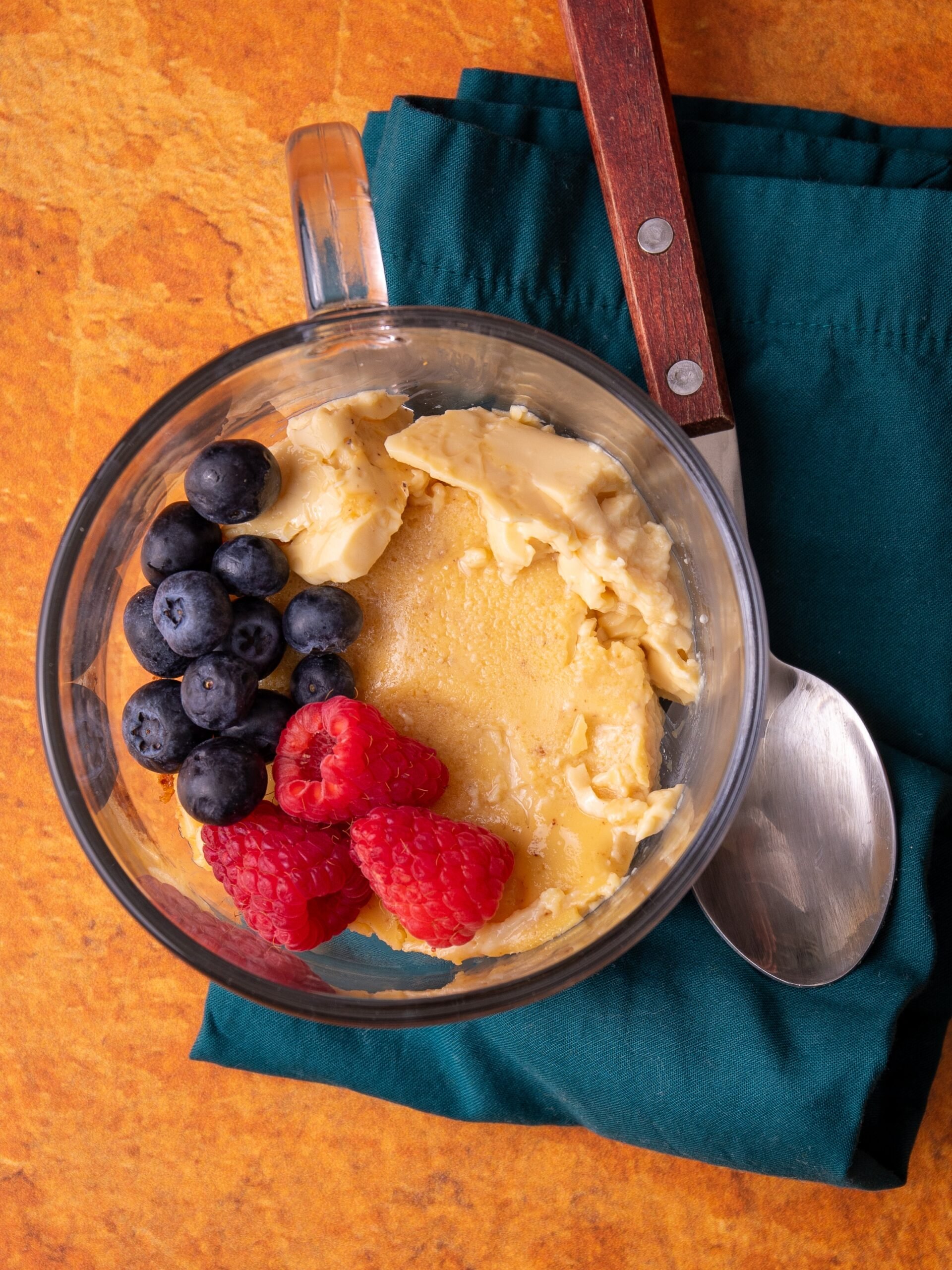 A glass cup of baked custard with fresh blue berries and raspberries. A silver spoon with a wooden handle is on the right. The handle is pointing up to the top and the spoon is resting on a green napkin. The entire image in on an orange textured background. 