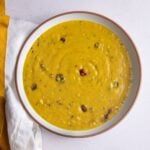 Overhead bowl of Guyanese yellow split pea dhal with a wiri wiri pepper, in a white bowl with a terracotta rim beside a mustard linen napkin