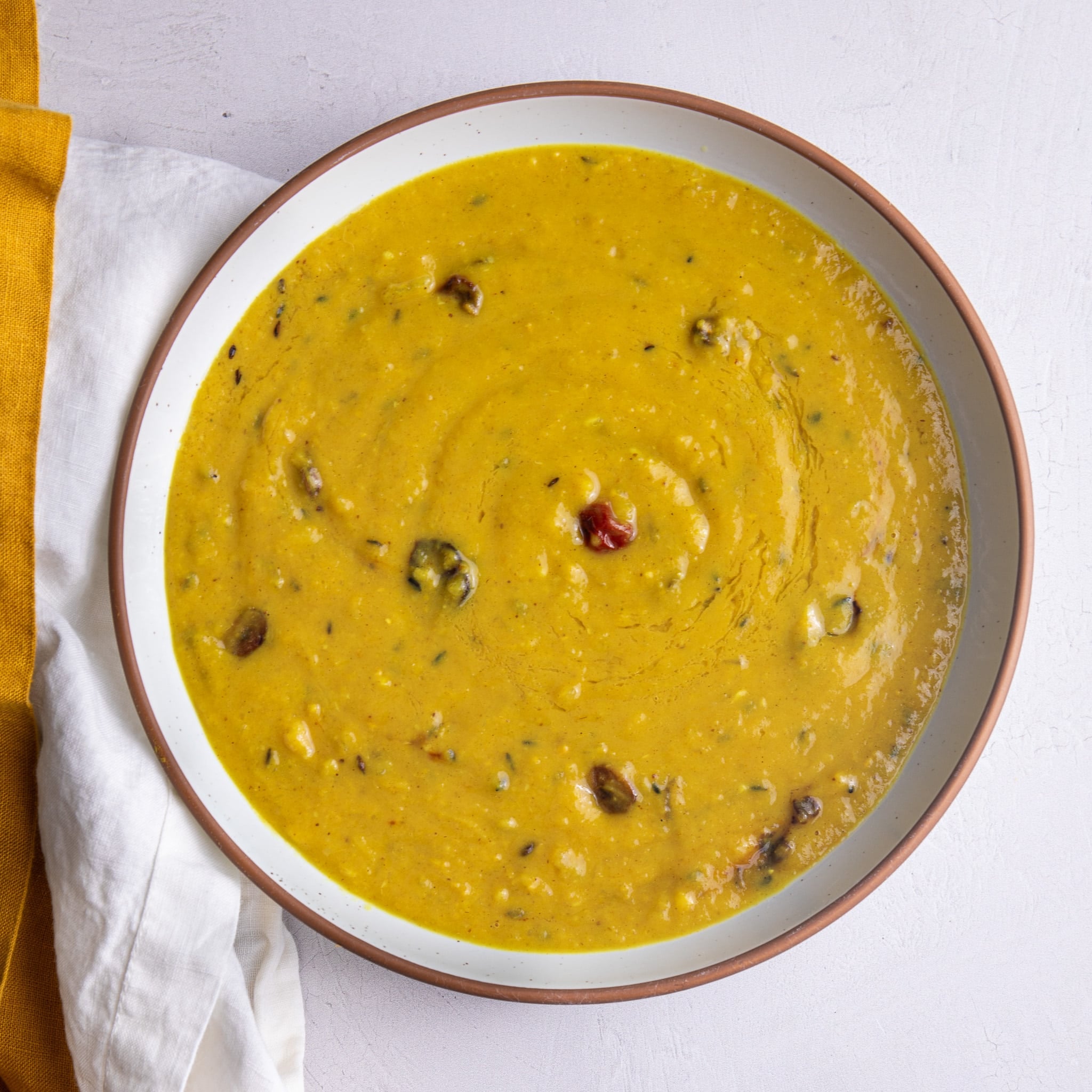 Overhead bowl of Guyanese yellow split pea dhal with a wiri wiri pepper, in a white bowl with a terracotta rim beside a mustard linen napkin