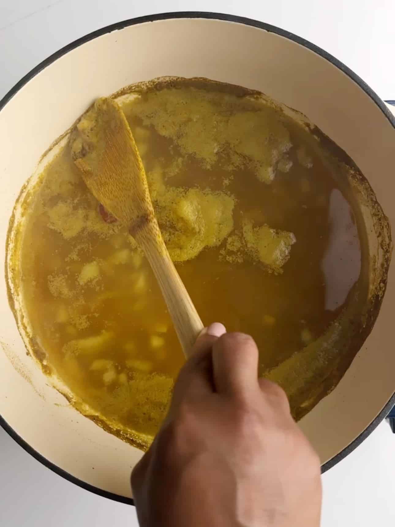 A brown hand holding a wooden spoon is skimming some of the scum off the top of the boiling dhal