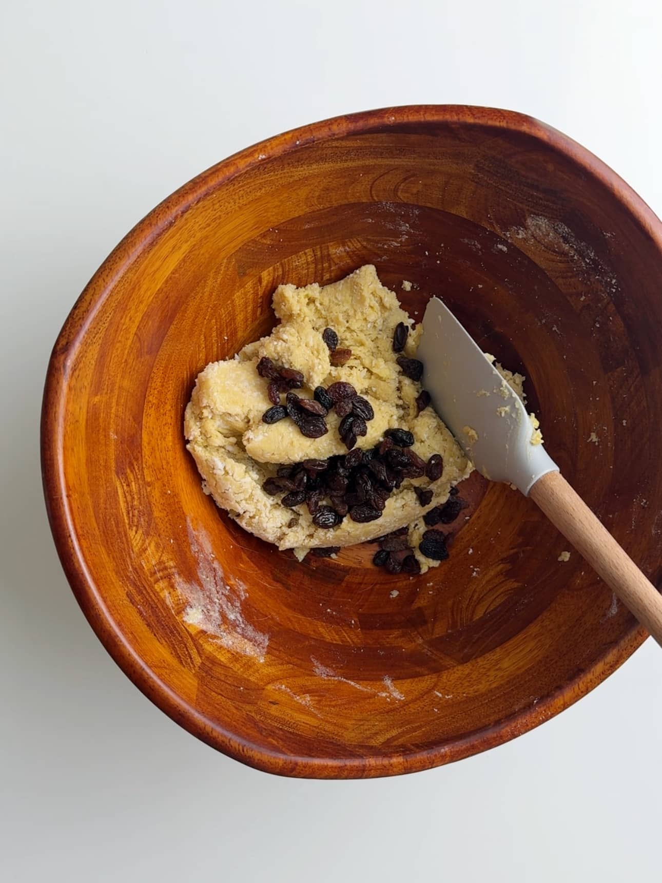 Adding raisins to a bowl of coconut buns dough. The bowl is wooden and sitting on a white surface with a gray silicons spatula with a wooden handle resting in the bowl.