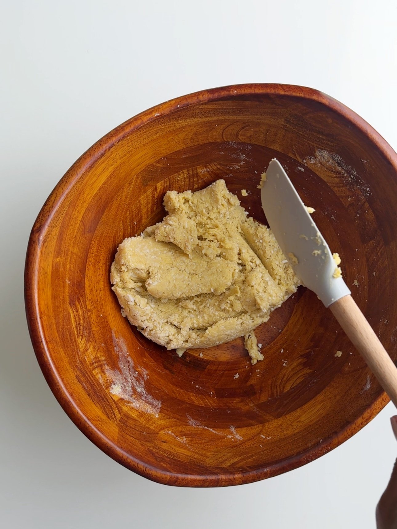 Dough for coconut buns in a brown wooden bowl with a gray silicone spatula in the bowl