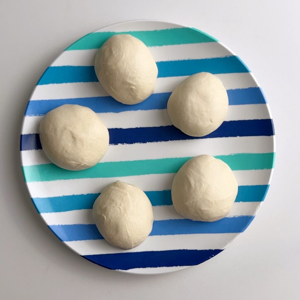 Five smooth white dough balls on a multi striped plate. The plate has alternating navy, sky blue, blue and teal stripes on a white background