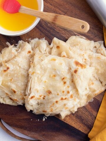 Guyanese paratha oil rotis folded on a round wooden board. A bowl with oil is visible on the left and a metal rolling pin is visible on the right