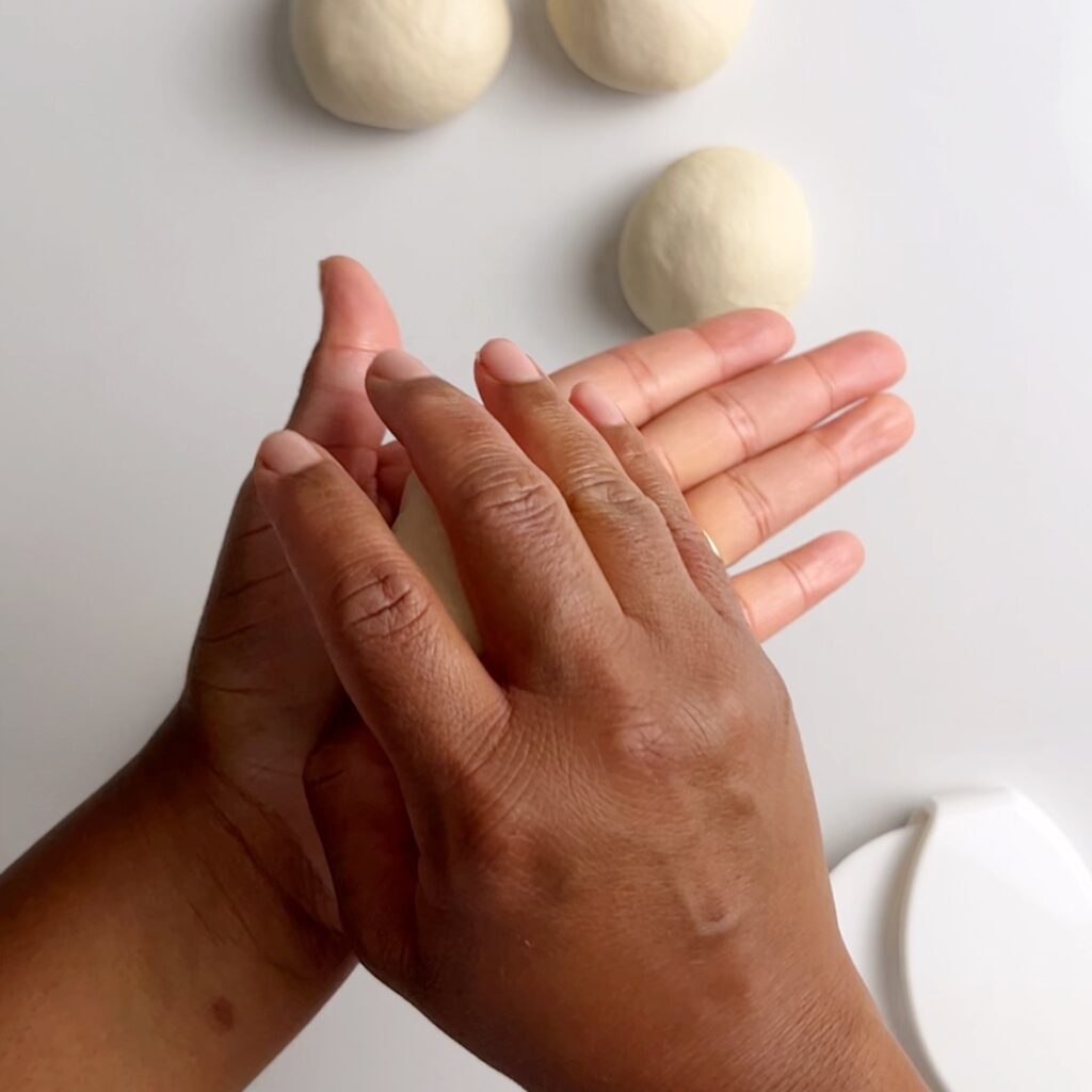 Brown hands holding dough. There are three smooth roti dough balls visible in the back ground. They are resting on a white surface with a white dough scraper in the lower right of the image