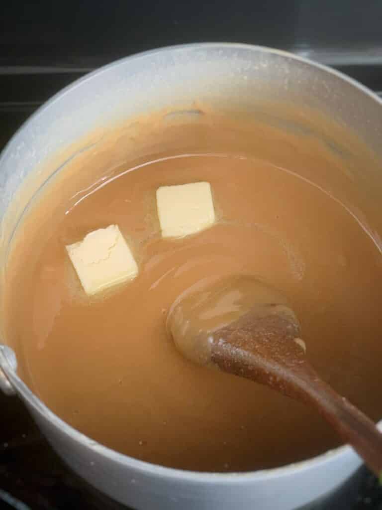 Fudge in a sauce pan with a wooden spoon in the mixture and two pats of butter on top of the fudge