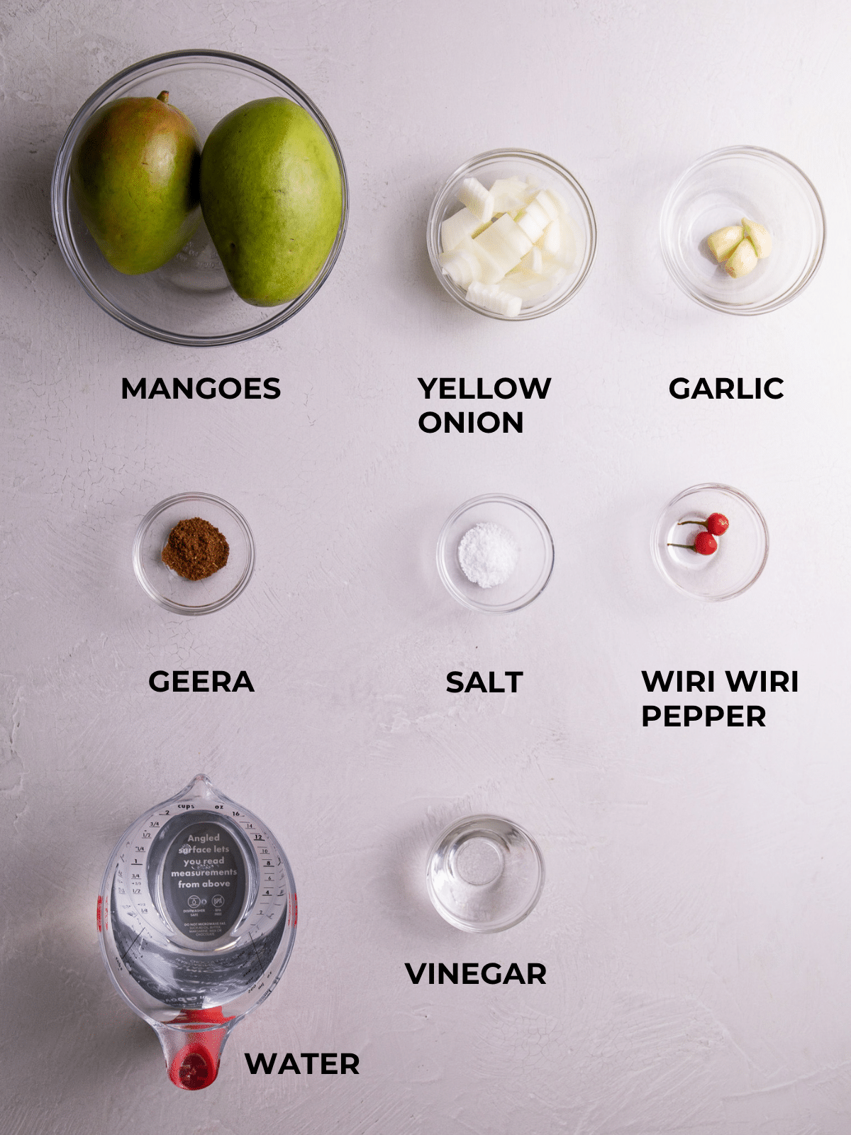 Flat lay of ingredients for Guyanese mango sour including green mangoes, yellow onion, garlic, geera, salt, wiri wiri pepper, vinegar, and water.