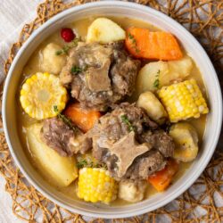 Bowl of Caribbean oxtail soup with tender oxtail, corn on the cob, dumplings, cassava, plantain, and sweet potato in a golden split pea broth, garnished with thyme and a wiri wiri pepper