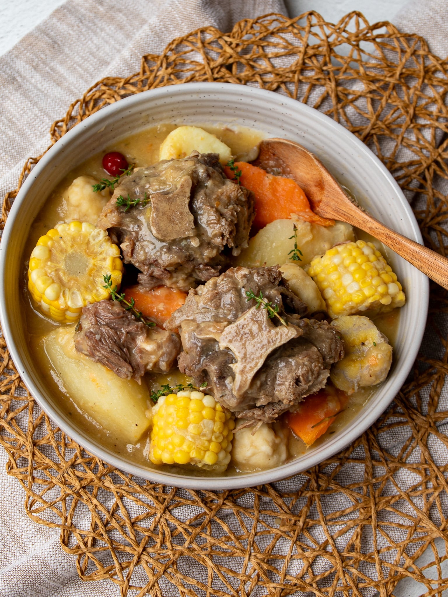 Angled view of Caribbean oxtail soup with oxtail, corn, dumplings, sweet potato, and a wiri wiri pepper in a cream bowl with a wooden spoon