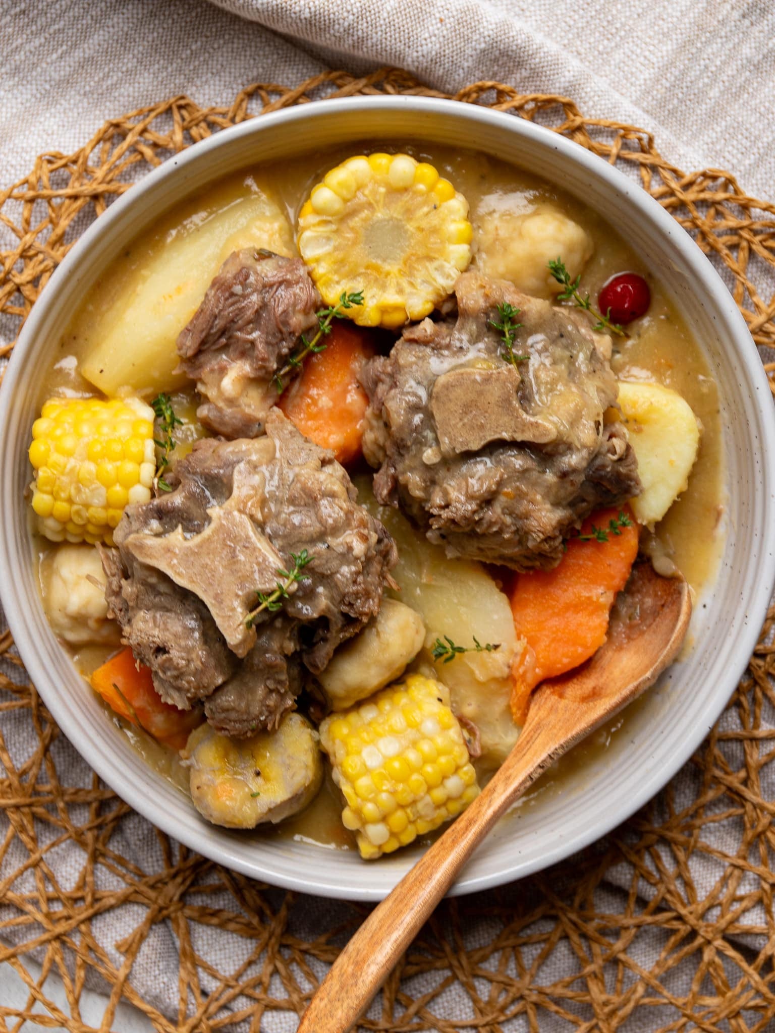 Bowl of Caribbean oxtail soup with a wooden spoon, showing tender oxtail with the bone, corn on the cob, dumplings, cassava, and sweet potato in a golden broth