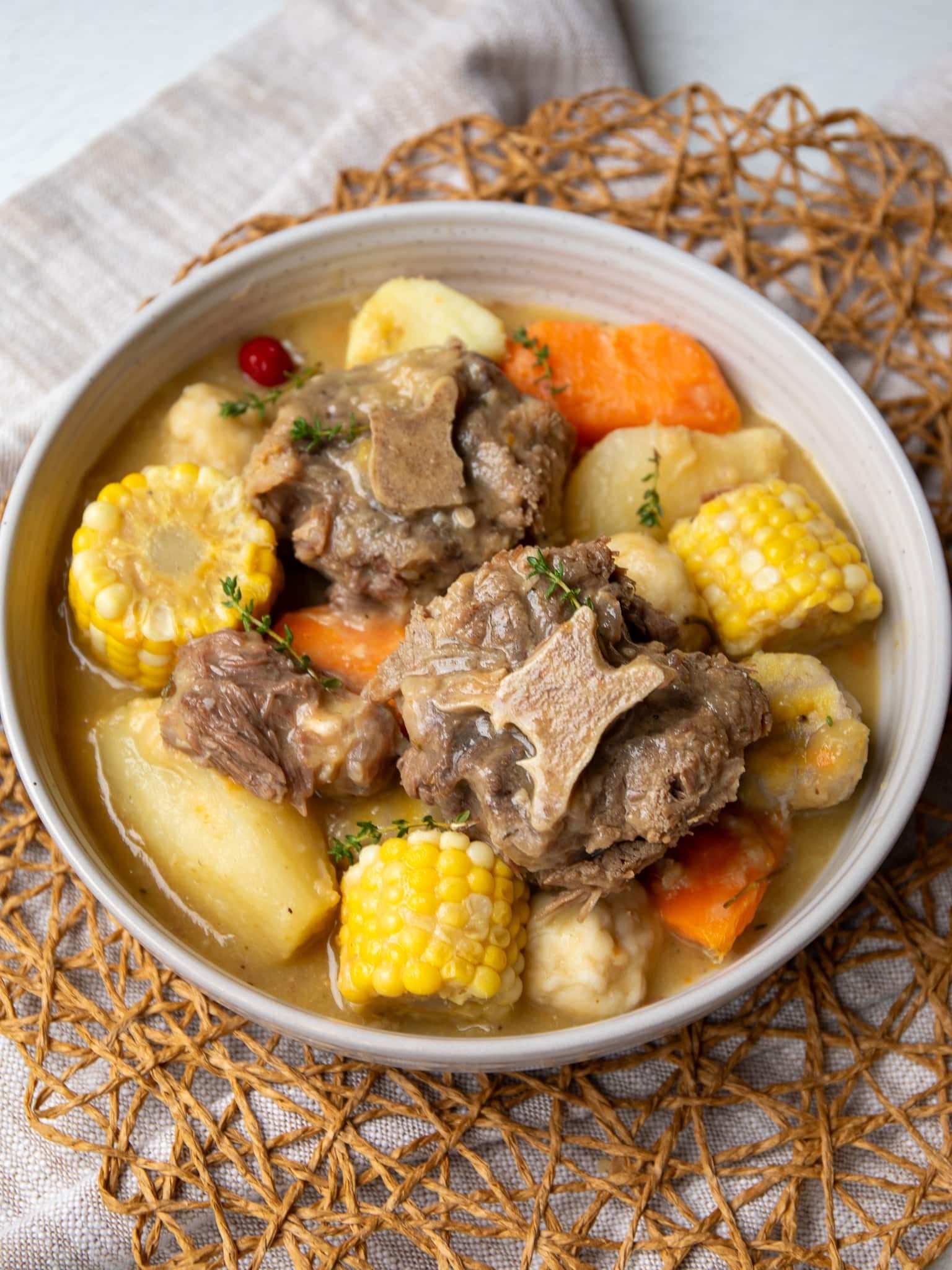 Bowl of Caribbean oxtail soup with tender oxtail, corn on the cob, dumplings, cassava, plantain, and sweet potato in a golden split pea broth, garnished with thyme and a wiri wiri pepper