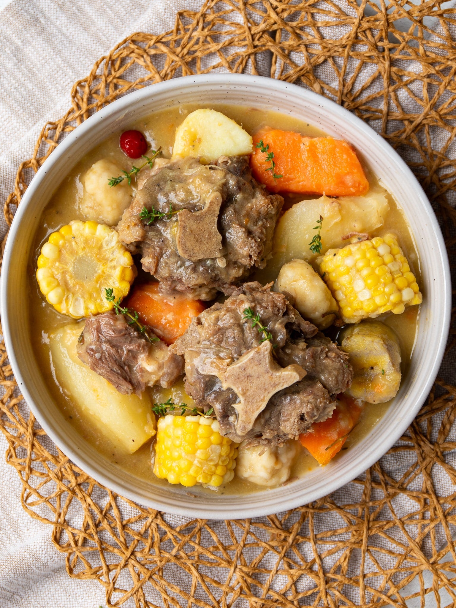 Bowl of Caribbean oxtail soup with tender oxtail, corn on the cob, dumplings, cassava, plantain, and sweet potato in a golden split pea broth, garnished with thyme and a wiri wiri pepper