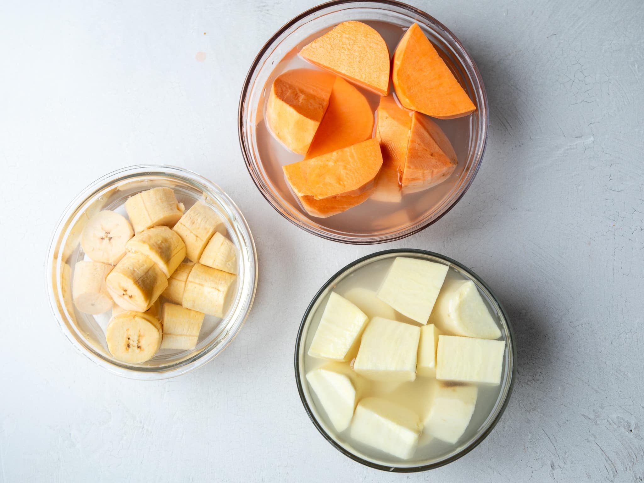 Cut sweet potatoes, white sweet potatoes, and plantains soaking in bowls of water to keep them from browning