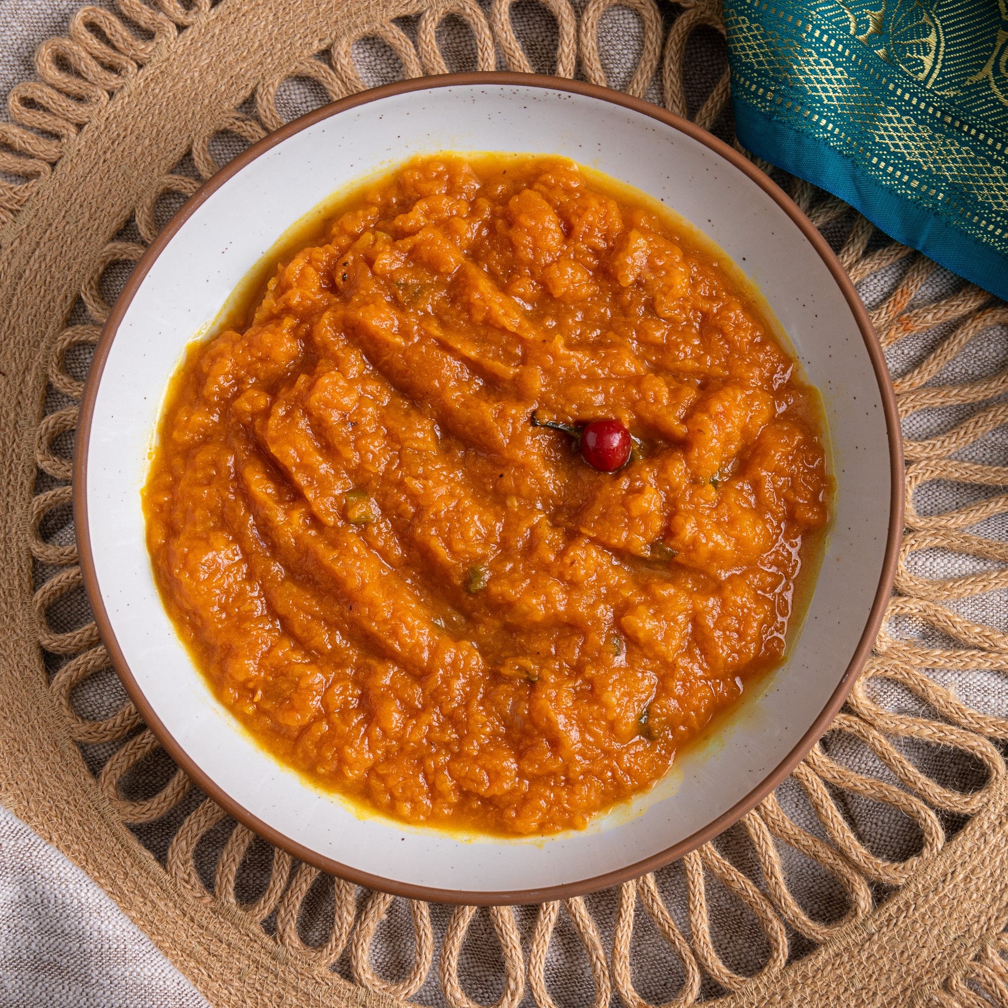 Overhead bowl of Guyanese sautéed pumpkin with a wiri wiri pepper, on a woven placemat with a teal sari
