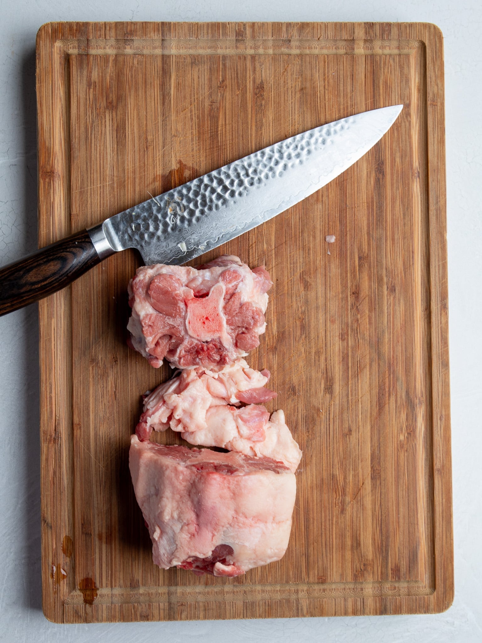Oxtail on a wooden cutting board showing a trimmed piece, the thick layer of fat removed, and an untrimmed piece, with a chef’s knife