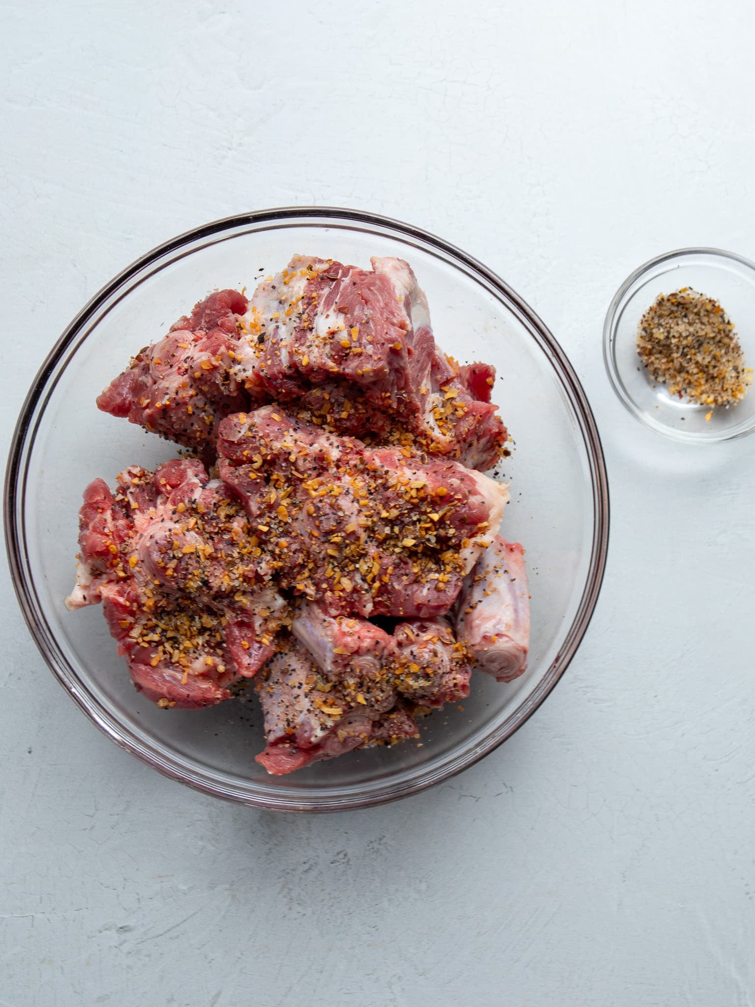 Raw oxtail pieces seasoned with steak seasoning in a glass bowl, with a small dish of extra seasoning on the side