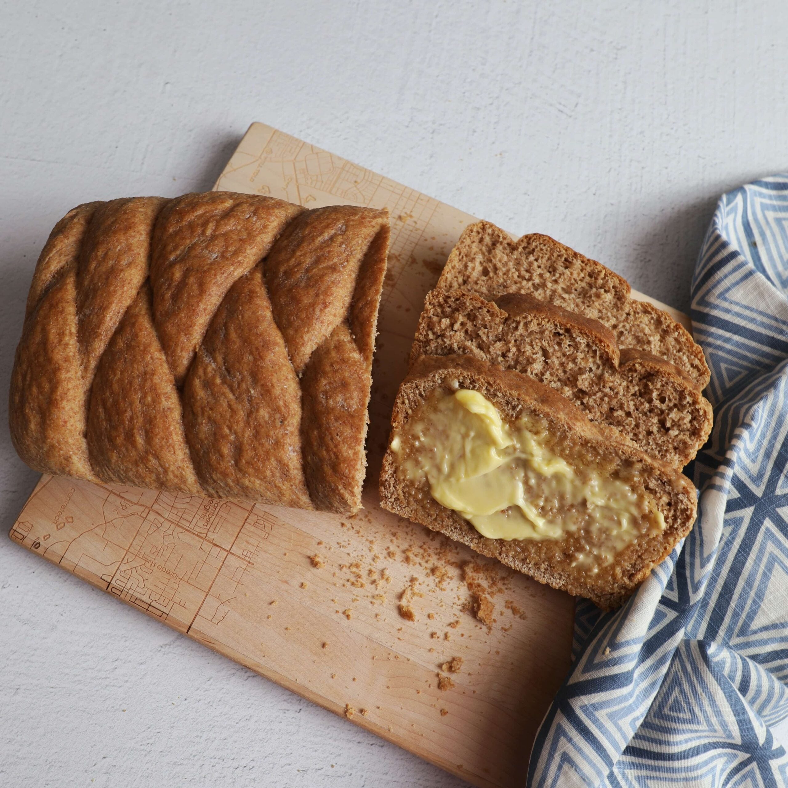 whole wheat loaf of bread on a bread board with three slices. The slice closest to the front is spread with butter. There is a white napkin with a blue triangular pattern to the right of the bread slices.