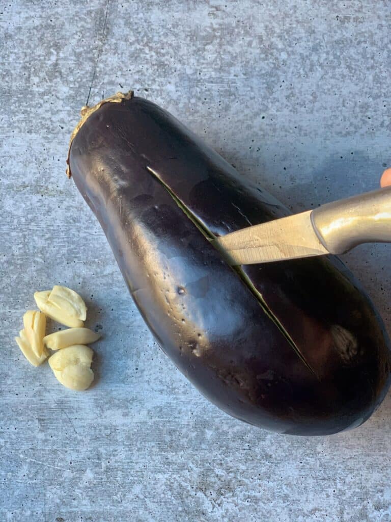 a knife inserted in a ripe eggplant that's been sliced down middle with halved garlic cloves to the side