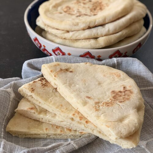 Sada rotis cut in half placed on a grey napkin with a bowl of fluffy sada rotis in the back.