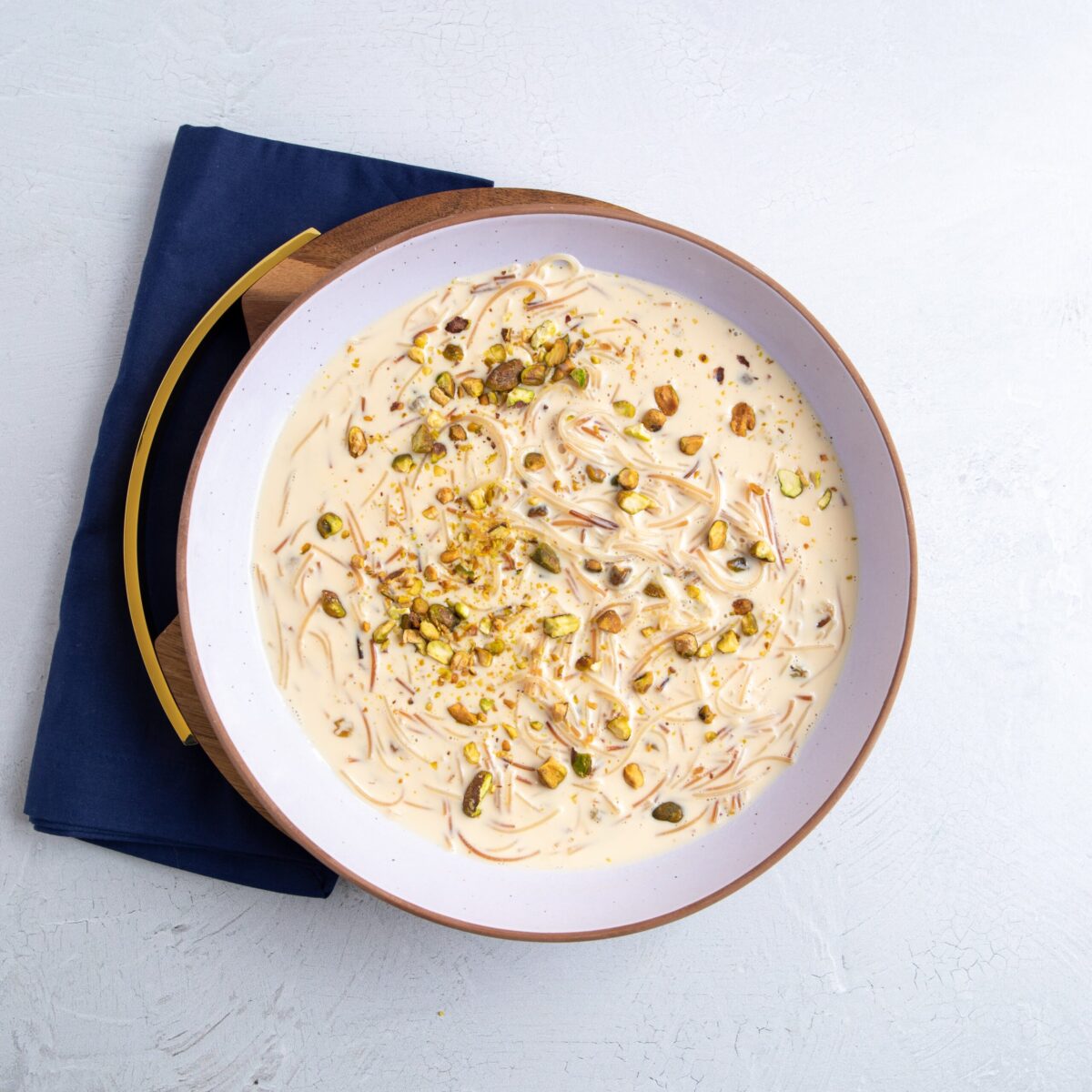 vermicelli noodles in a creamy base in a white bowl with brown border, resting on a round wooden board with a gold border. Everything is resting on a blue napkin folded into a rectangle.