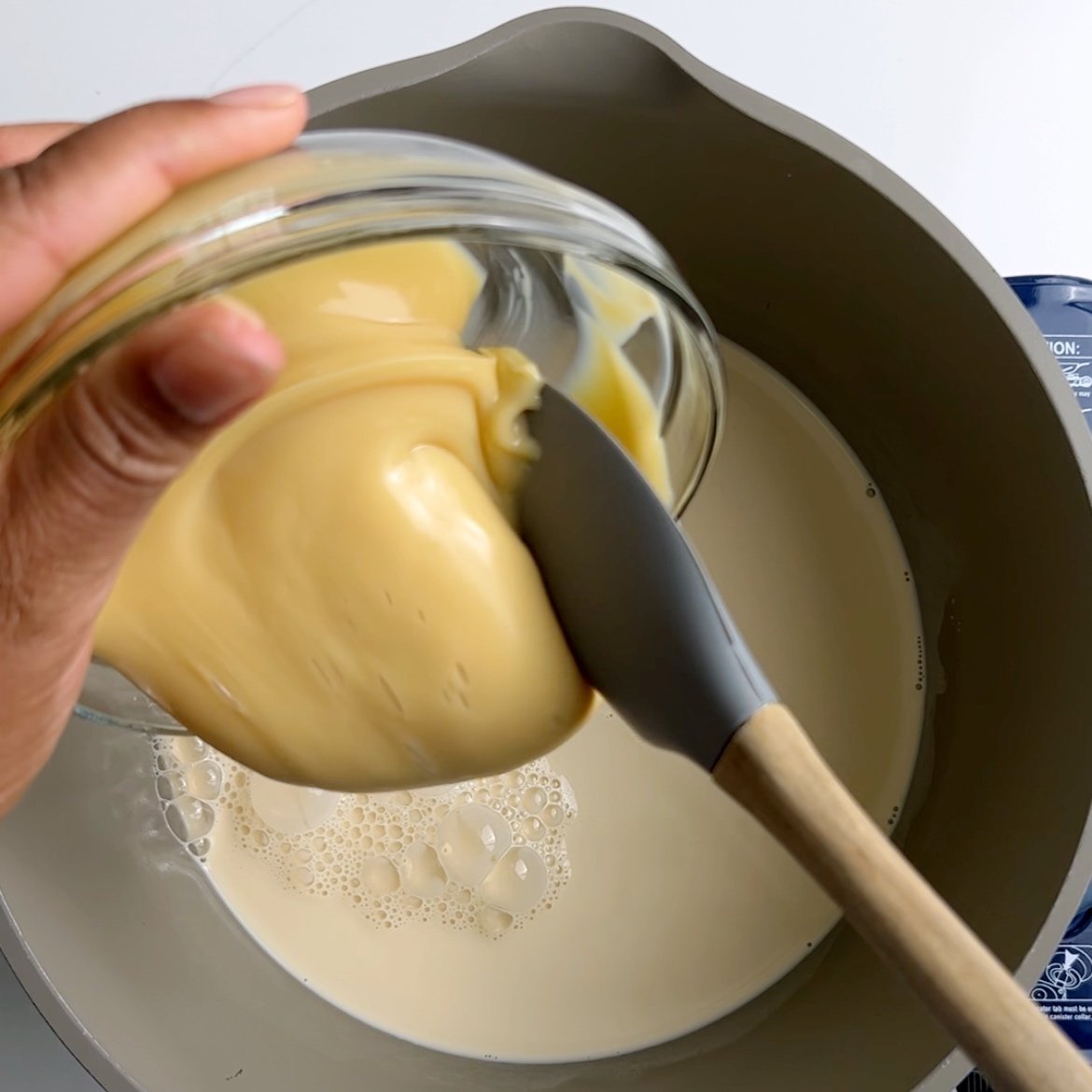 Scooping Condensed milk with a rubber spatula, into a grey stock pot of milk