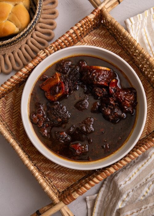 Guyanese Pepperpot in a bowl sitting in a hand woven indigenous sifter. To the left a piece of plait bread is visible and to the bottom right there is a cream and white stripped napkin with gold thread running through the stripes.