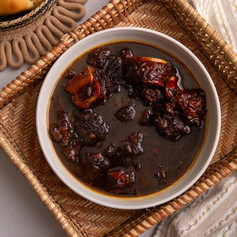 Guyanese Pepperpot in a bowl sitting in a hand woven indigenous sifter. To the left a piece of plait bread is visible and to the bottom right there is a cream and white stripped napkin with gold thread running through the stripes.