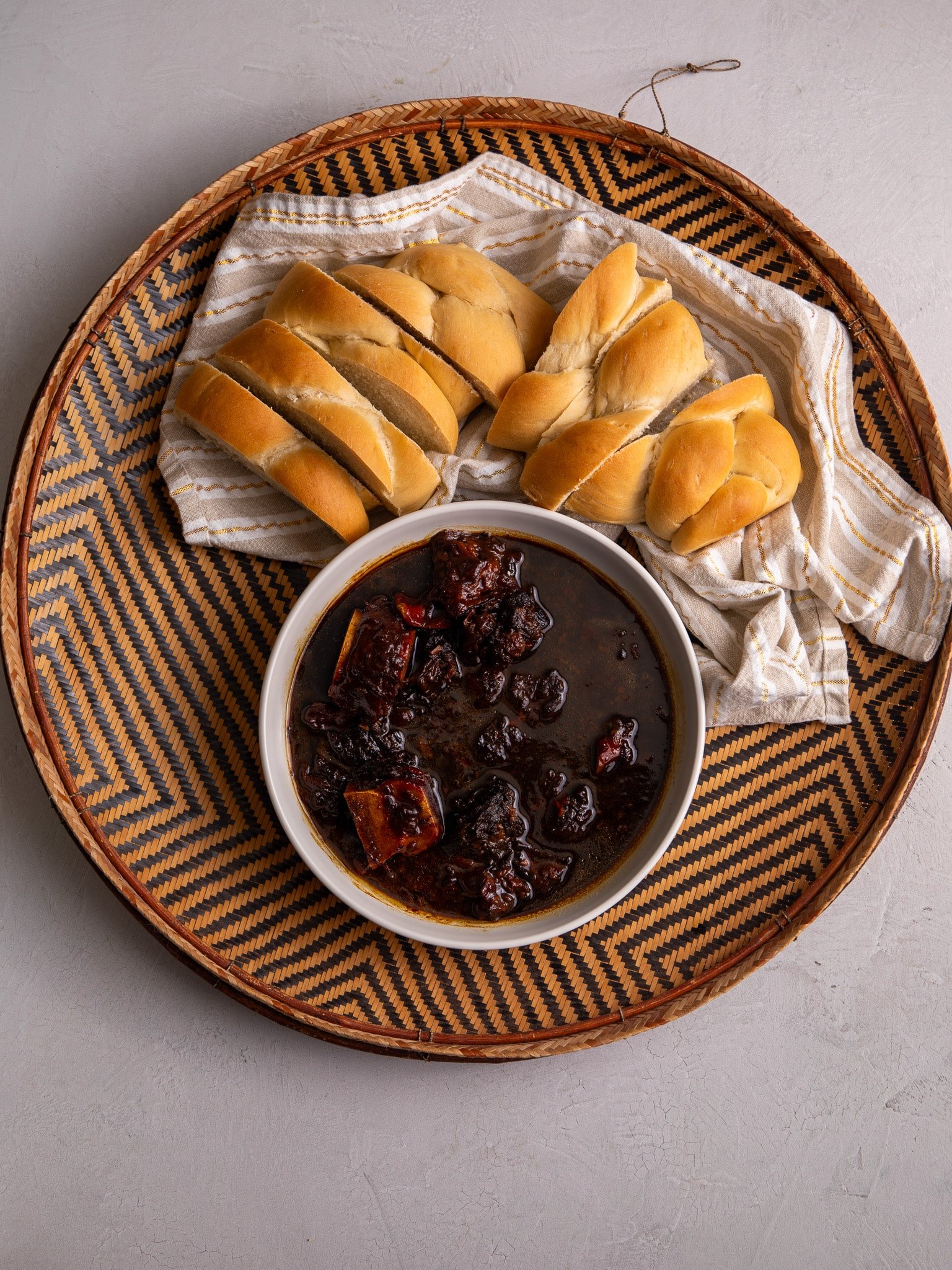 Guyanese Pepperpot in a large round woven basket.The Pepperpot is in a cream ceramic bowl. Above the bowl there is sliced up plait bread on a a napkin. 