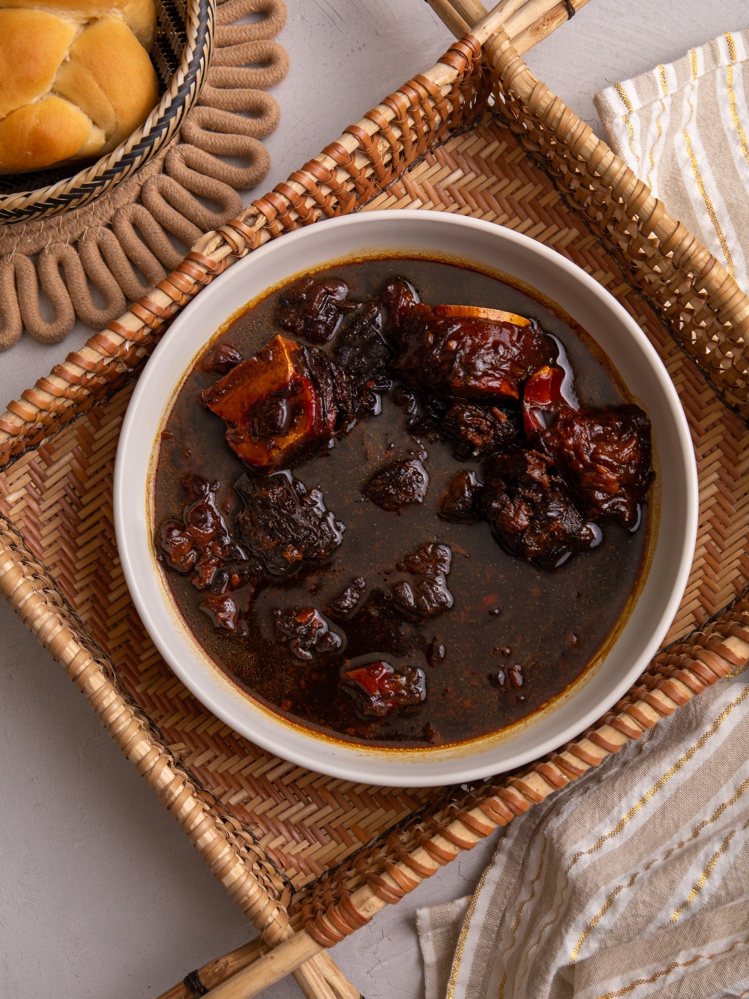 Guyanese Pepperpot in a bowl sitting in a hand woven indigenous sifter. To the left a piece of plait bread is visible and to the bottom right there is a cream and white stripped napkin with gold thread running through the stripes.