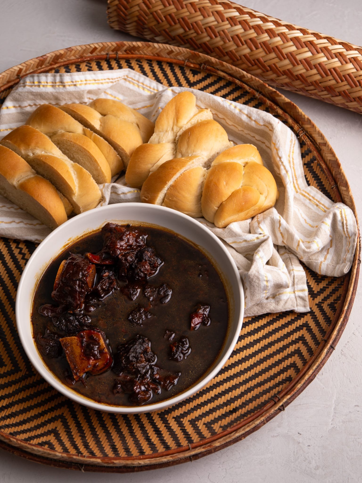 Guyanese Pepperpot in a bowl sitting in a hand woven indigenous sifter. To the left a piece of plait bread is visible and to the bottom right there is a cream and white stripped napkin with gold thread running through the stripes.