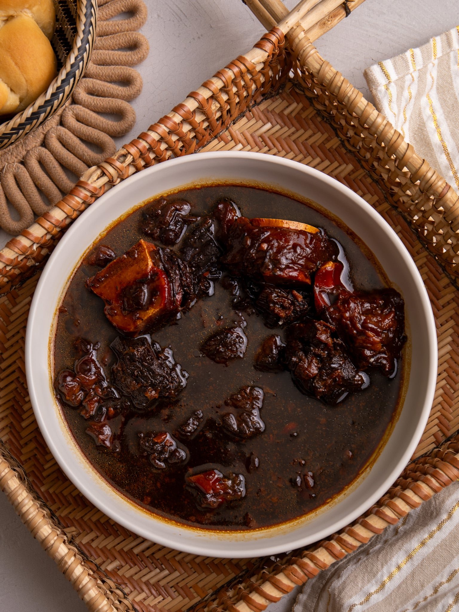 Guyanese Pepperpot in a bowl sitting in a hand woven indigenous sifter. To the left a piece of plait bread is visible and to the bottom right there is a cream and white stripped napkin with gold thread running through the stripes.