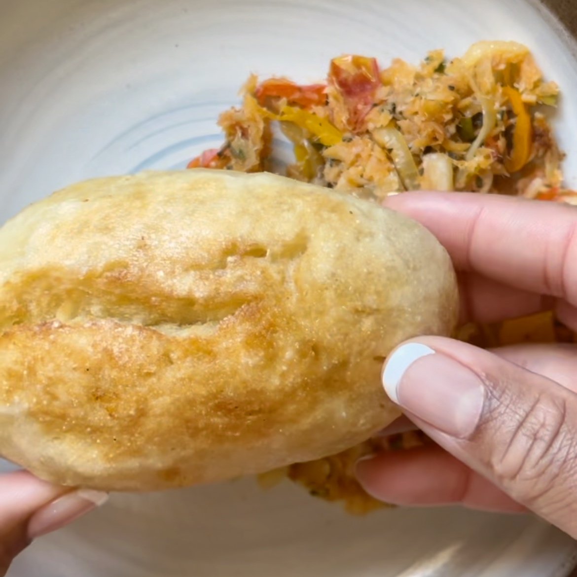 Fingers holding a golden brown fried duff over a plate of saltfish