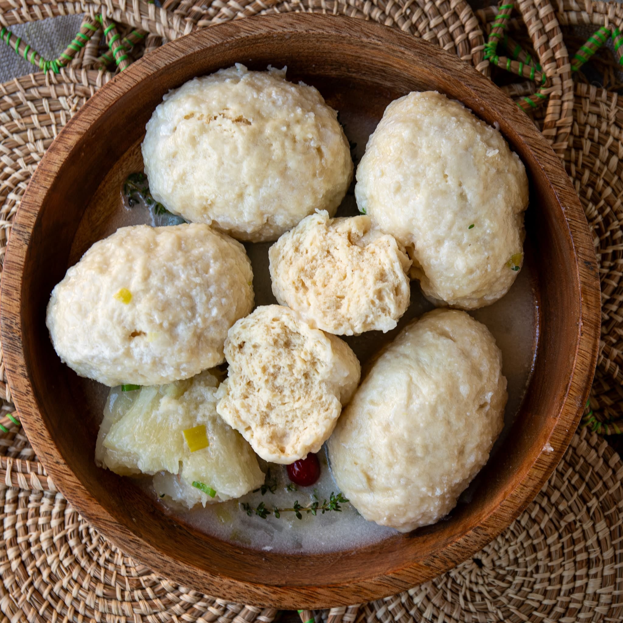 Overhead view of five Guyanese duff in a wooden bowl of coconut milk broth with cassava, thyme, scallions, and a wiri wiri pepper, one duff torn open to show its soft, fluffy inside.