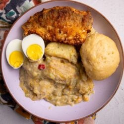 Close-up of a full plate of metem with mashed root vegetables, a whole dumpling, fried fish, and two halved boiled eggs on a floral napkin.