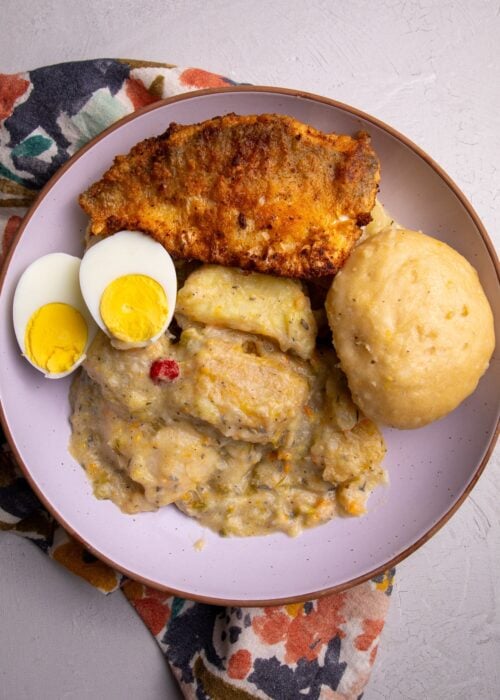 Close-up of a full plate of metem with mashed root vegetables, a whole dumpling, fried fish, and two halved boiled eggs on a floral napkin.