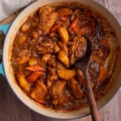 Guyanese chicken stew in a large dutch oven with a wooden spoon scooping some of the stew. A white kitchen towel is to the top left and the pot is sitting on a brown wooden table.