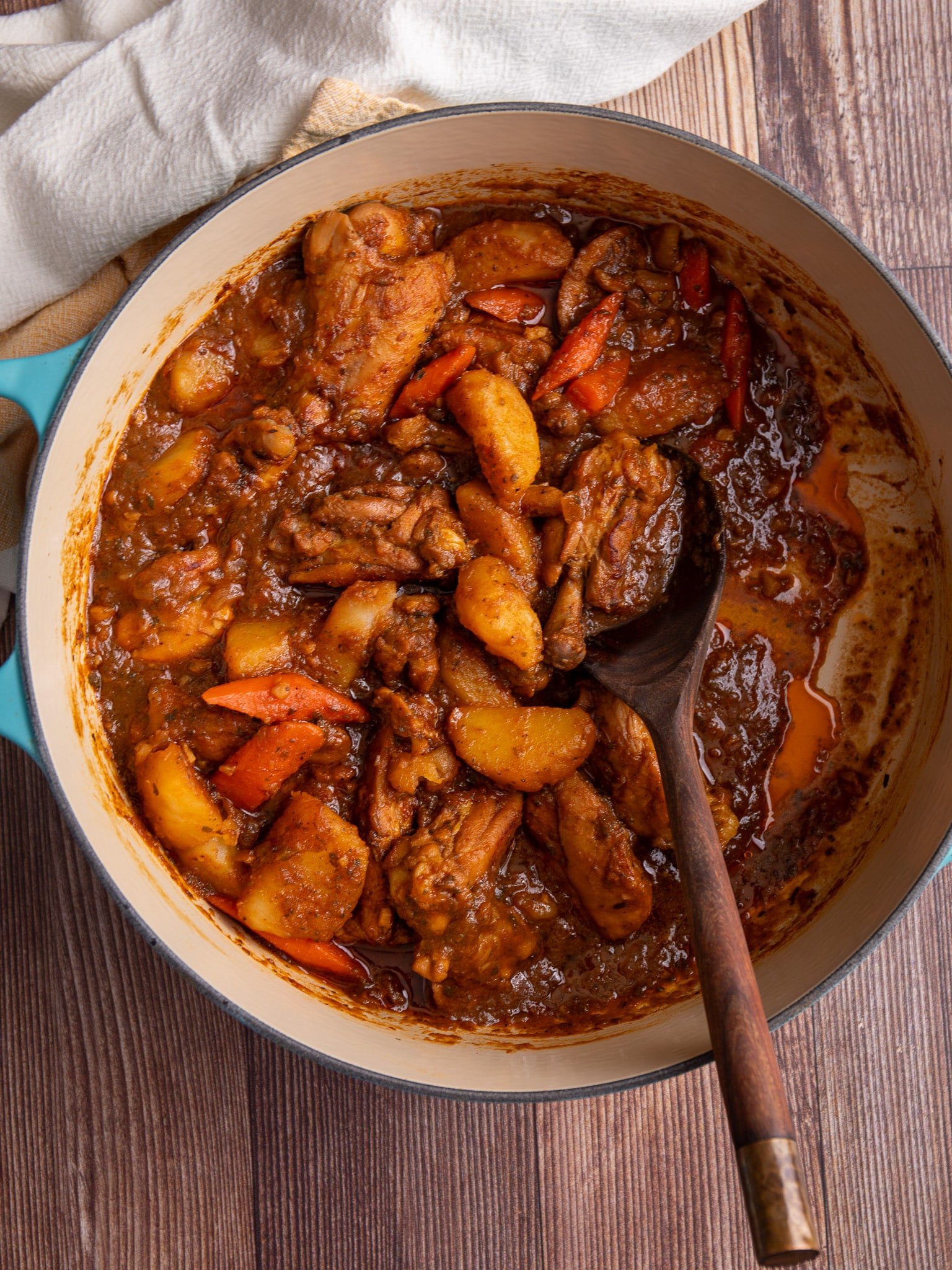 Guyanese chicken stew in a large dutch oven with a wooden spoon scooping some of the stew. A white kitchen towel is to the top left and the pot is sitting on a brown wooden table.