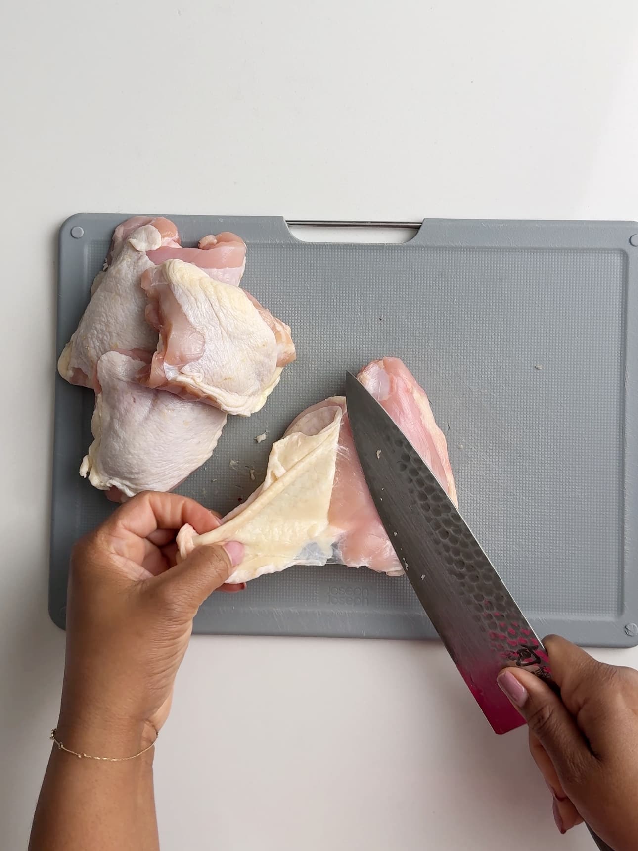 Chicken thighs are on a grey plastic cutting board on a white counter. Brown hands are holding a chef's knife and removing the skin from the chicken thighs. The left hand is pulling the skin and the right hand is holding the knife that is pressed against the chicken thigh.
