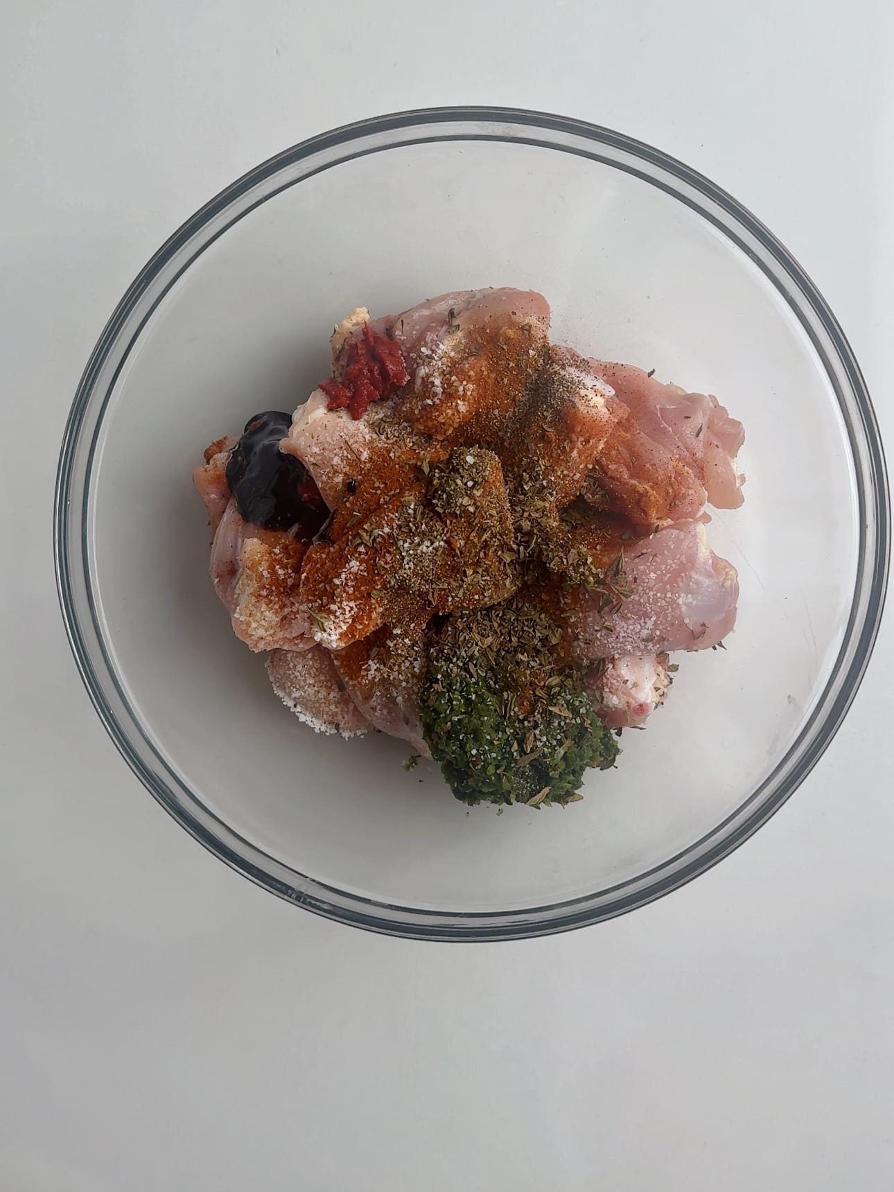 Chicken thighs and seasoning in a glass mixing bowl on a white counter. 