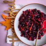 cranberry chutney in a white plate with a gold spoon taking a scoop. A carving of the word thankful is tucked under it and on top of a white napkin.