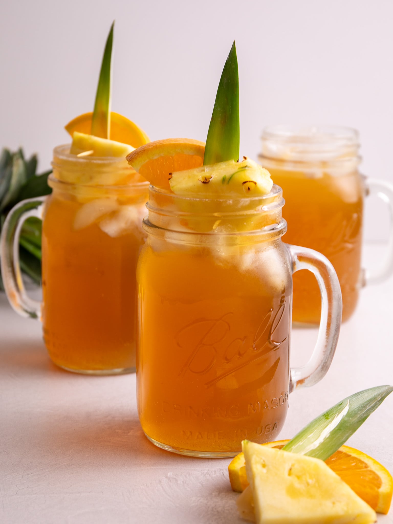 Mason jar mug of fermented pineapple drink garnished with pineapple, orange slice, and pineapple leaf, with two drinks in the background.