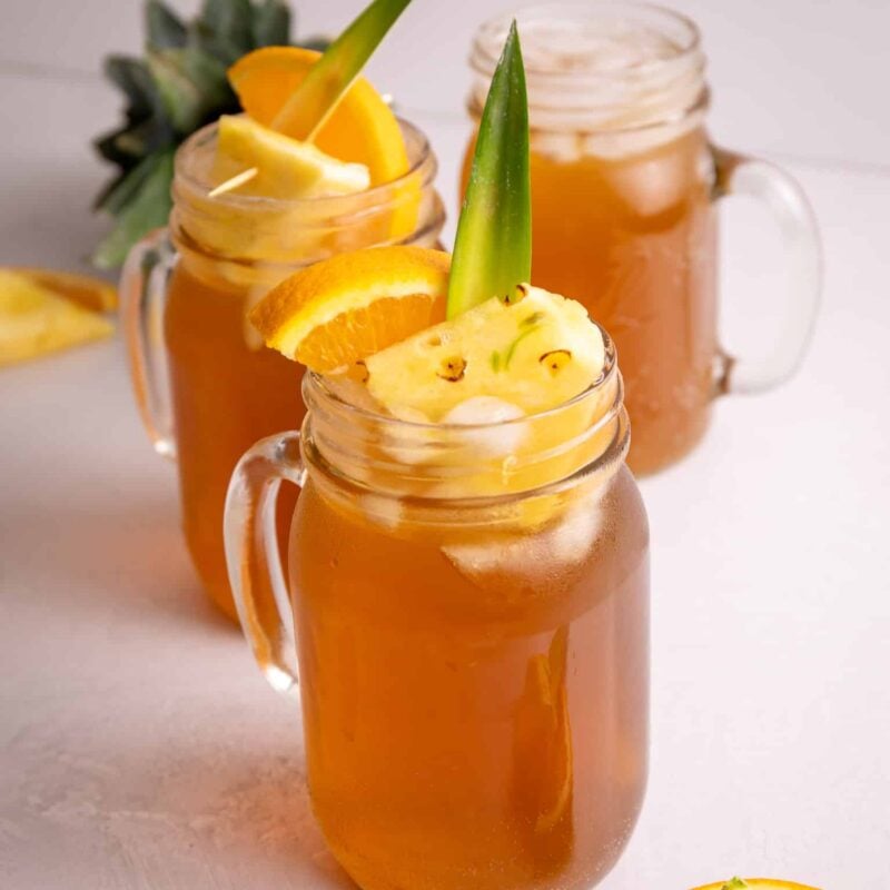 Three glasses of pineapple drink garnished with oranges and pineapples.