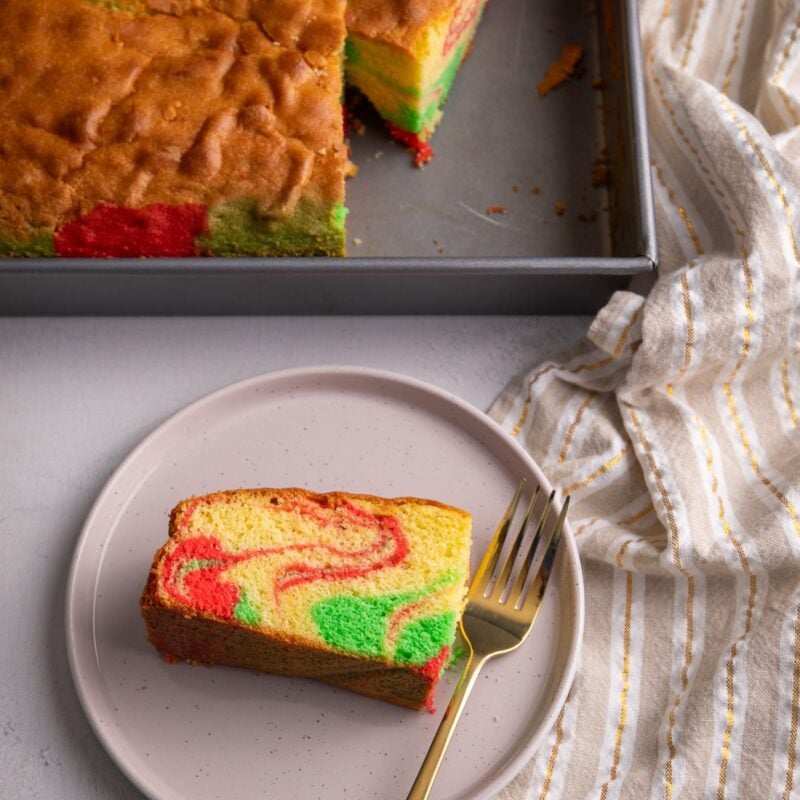 Guyanese Sponge Cake with red and green swirls on a cream circular plate. In the background there is a rectangular pan with a slice of cake and a cream and gold striped towel to the side.