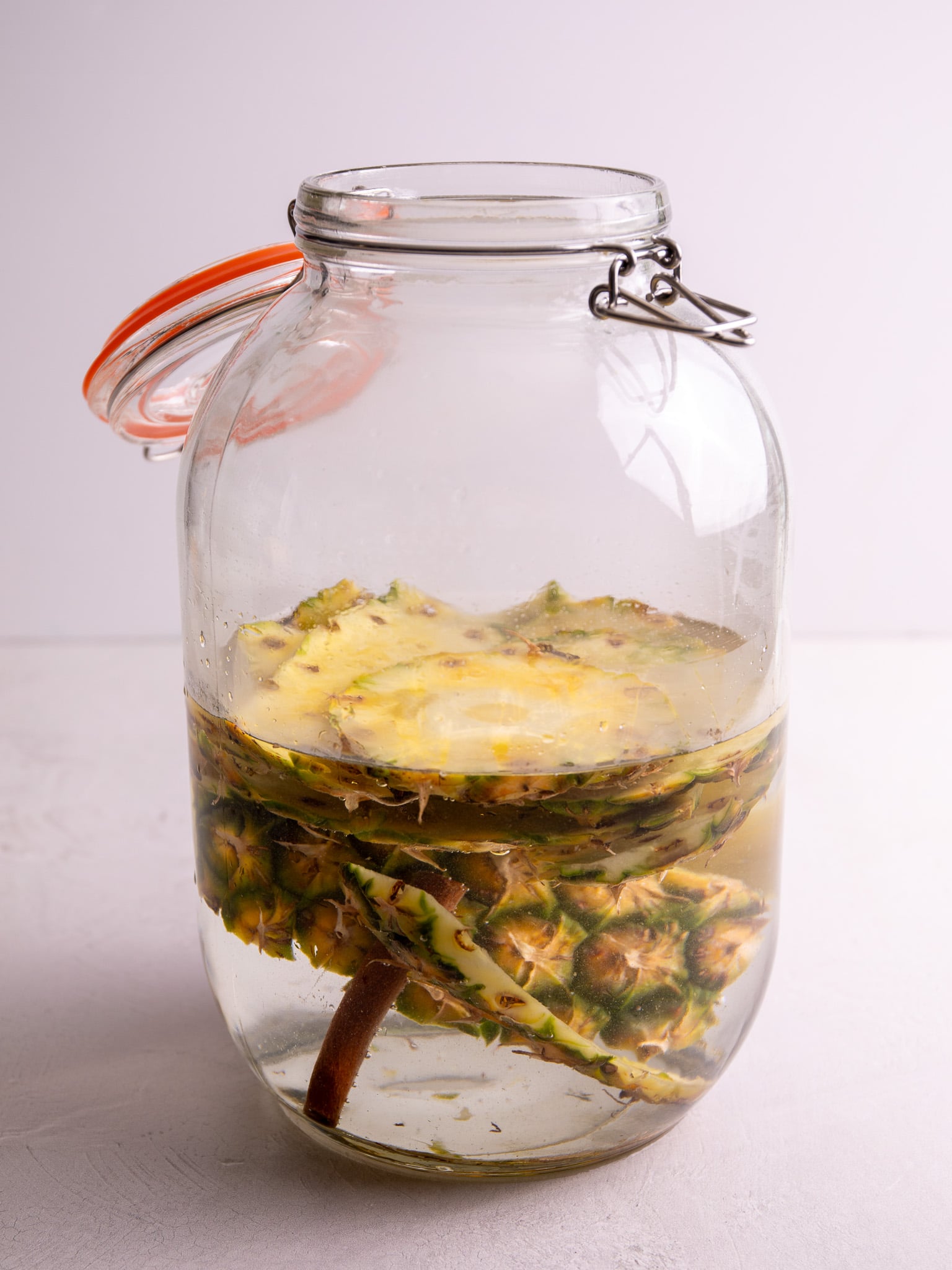 Glass fermentation jar with pineapple peels, cores, and cinnamon stick soaking in liquid.