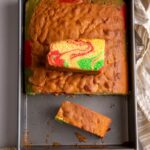 Sponge cake in a rectangular pan. The cake is cut with about a third missing. A slice is resting in that space and another slice is sitting on top of the cake to reveal a cross section of red and green swirls.
