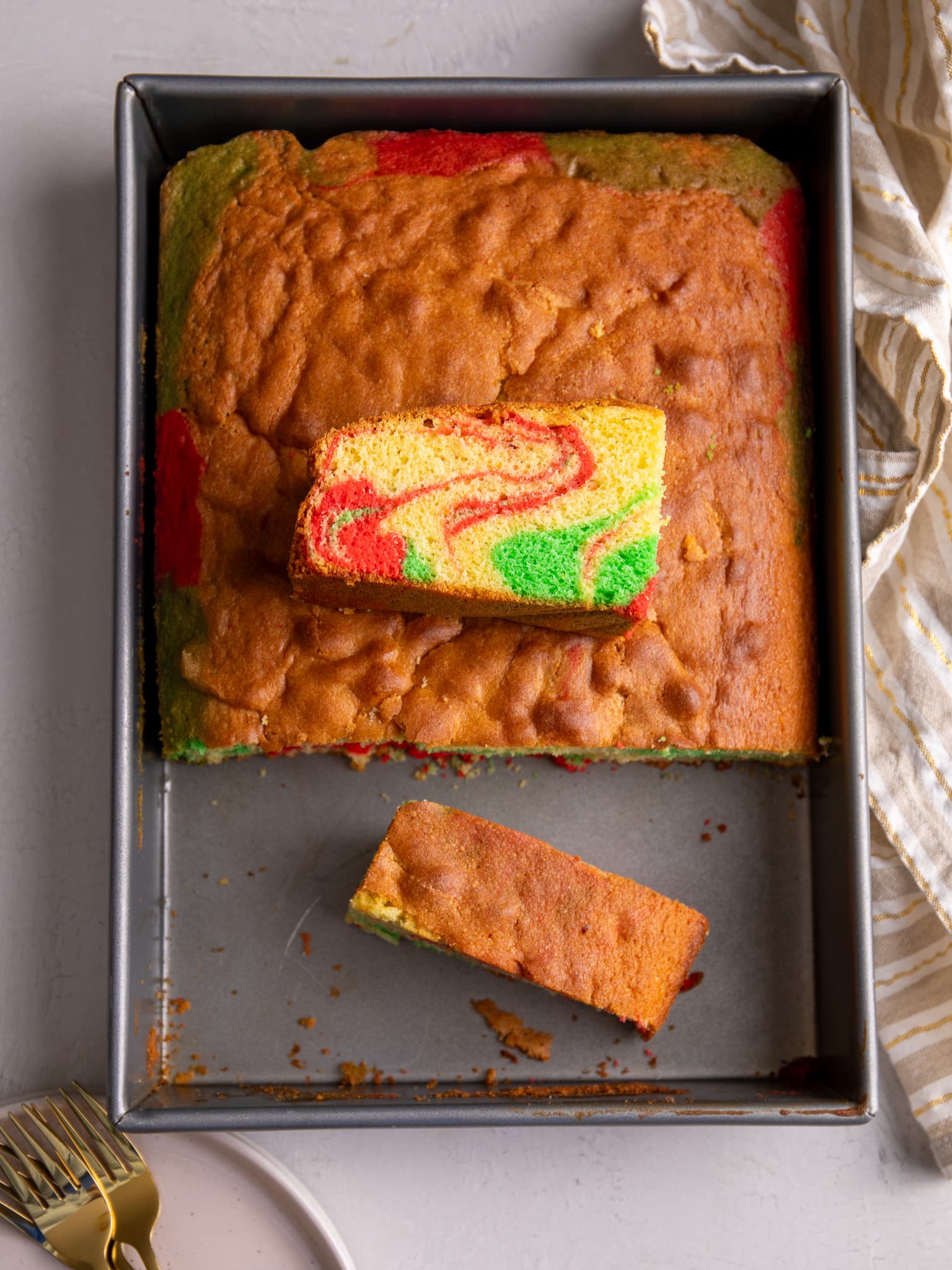 Sponge cake in a rectangular pan. The cake is cut with about a third missing. A slice is resting in that space and another slice is sitting on top of the cake to reveal a cross section of red and green swirls.